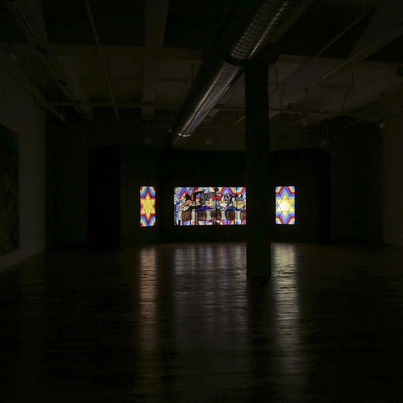 Color photograph of a glowing stained glass work at the back of a darkened gallery