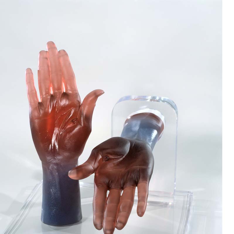Sculpture of two hands in earth tones on a clear plinth where one is reaching upward and the other is extending toward the viewer