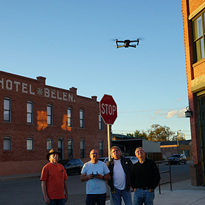 Color photograph of a drone pilot and three men observing a black drone in flight above a street corner with brick buildings in the background