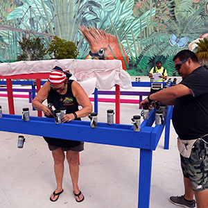 Color photograph of Mary Costa, Gabriel Garcia, and Rusty Johnson installing smoke and fireworks canisters on a rectangular, multicolored structure in a plaza with a large mural in the background