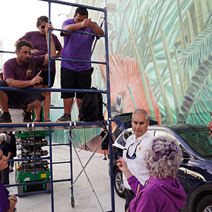 Color photograph of Judy Chicago and Chris Souza speaking to the Fire Marshall with Mary Costa, Gabriel Garcia, Vanessa Olvera, and Rusty Johnson around or on blue scaffolding with a car and a mural in the background