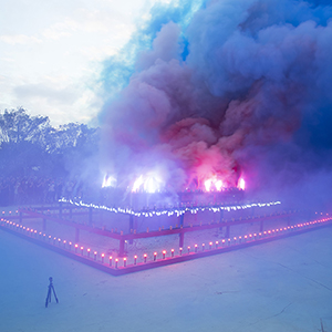 Color photograph of blue, purple, and pink smoke emerging from flares on a multicolored rectangular structure in a plaza as a crowd of people looks on