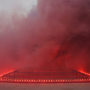 Color photograph of red and purple smoke emerging from flares on a multicolored rectangular structure in a plaza as a crowd of people looks on