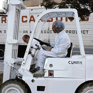 Color photograph of Lloyd Hamrol driving a white forklift as two people stand nearby