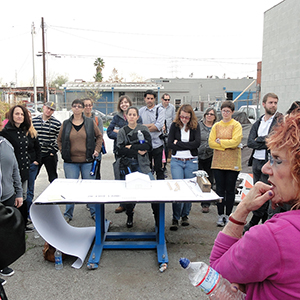 Color photograph of Glenn Phillips, Judy Chicago, and Jenna Didier speaking to a group of people standing around a table with a model on it