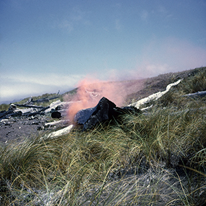 Color photograph of orange smoke rising behind a dark rock on a grassy hillside strewn with driftwood