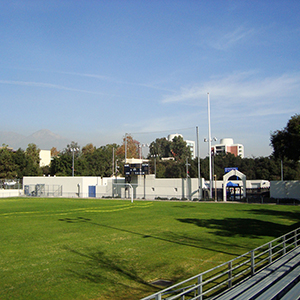 Color photograph of yellow caution tape strung across a sports field with a scoreboard and buildings in the background