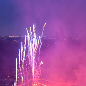 Color photograph of an aerial view of purple smoke and fireworks shooting up from a glowing red outline of a butterfly on a lawn in a park