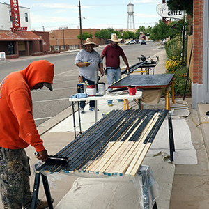 Color photograph of three people painting wood strips at tables set up on the sidewalk outside a brick building