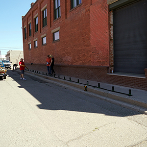 Color photograph of Jim Perry, Larry Scriptor, and Megan Schultz standing alongside a brick building with a row of smoke canisters installed on the curb