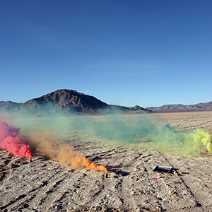 Color photograph of plumes of pink, orange, and yellow smoke emanating from the desert floor
