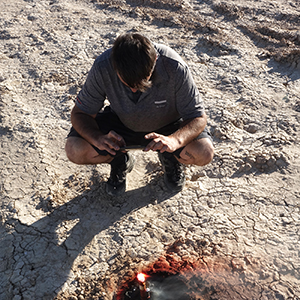 Color photograph of Chris Souza kneeling and taking a picture of a lit flare embedded in the desert floor