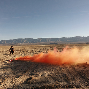Color photograph of Judy Chicago, standing, and Chris Souza, kneeling, in the desert looking at plumes of orange smoke