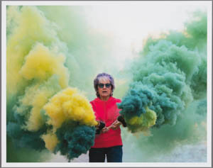 Print of Judy Chicago wearing a hot pink shirt and holding flares with green and yellow smoke emanating from them