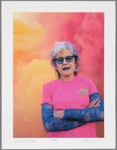 Print of Judy Chicago wearing a pink t-shirt with purple lace sleeves with arms crossed in front of a cloud of pink and orange smoke