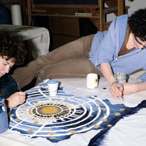 Volunteer Adrienne Weiss sits at a table applying blue paint onto a mock-up runner for Caroline Herschel while Judy Chicago lies across the table applying white paint onto the runner