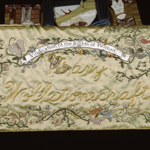 Bottom detail of the runner for Mary Wollstonecraft. Her name is in gold lettering surrounded by a design of flowers, vines, birds, butterflies, fish, and a white banner above with the text “Vindication of the Rights of Woman.”