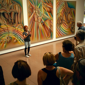Color photograph of Judy Chicago standing and talking to a group of people in front of three large, rainbow-colored paintings
