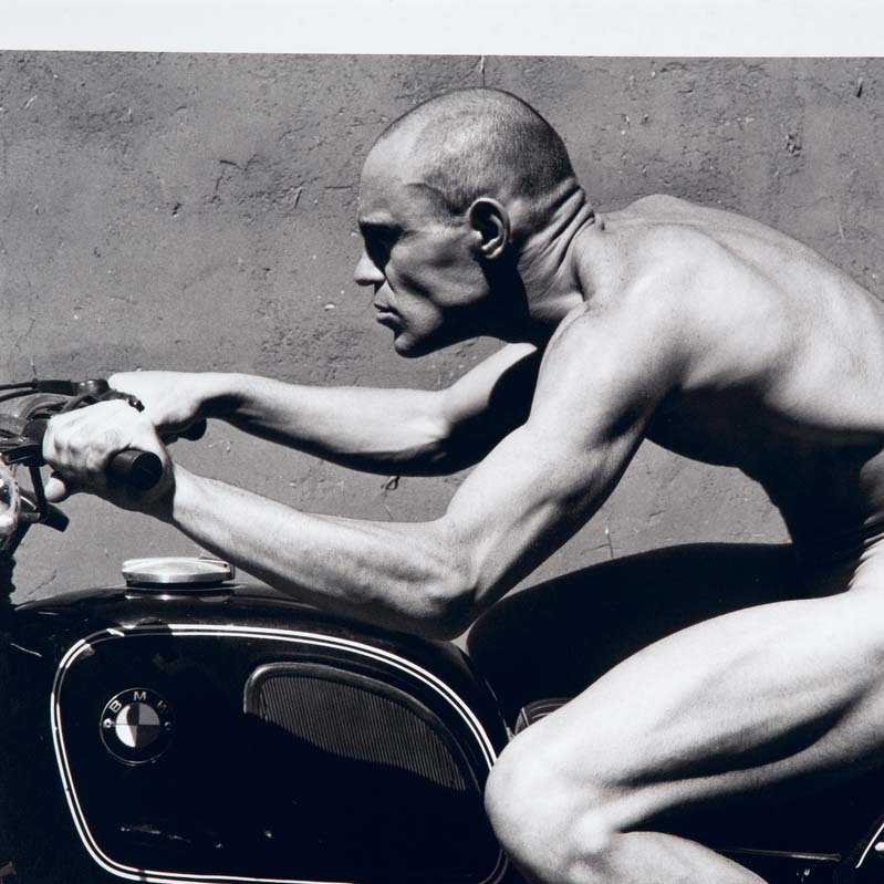 Black-and-white photograph of a nude man riding a motorcycle in profile