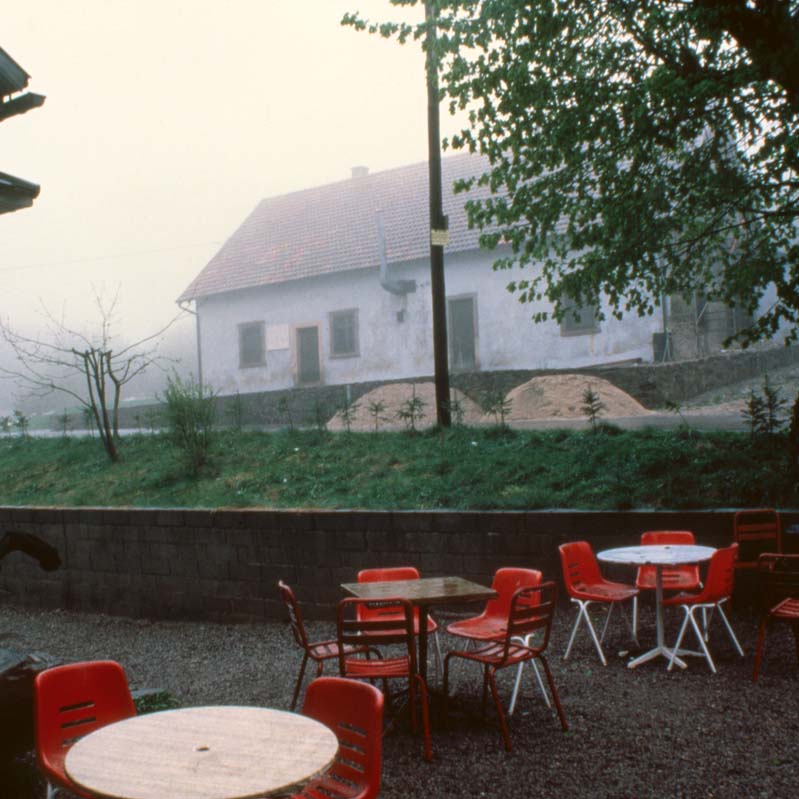 Color photograph of red chairs and brown tables outside a white building with another white building in the background
