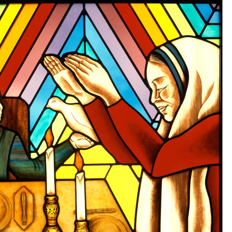 Detail of a stained glass work with a woman praying over a table where two people sit with a bird and candles