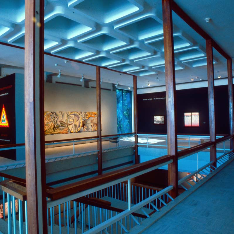 Color photograph of artworks installed on white and black walls in a second-story space with railings and a stairway in the center