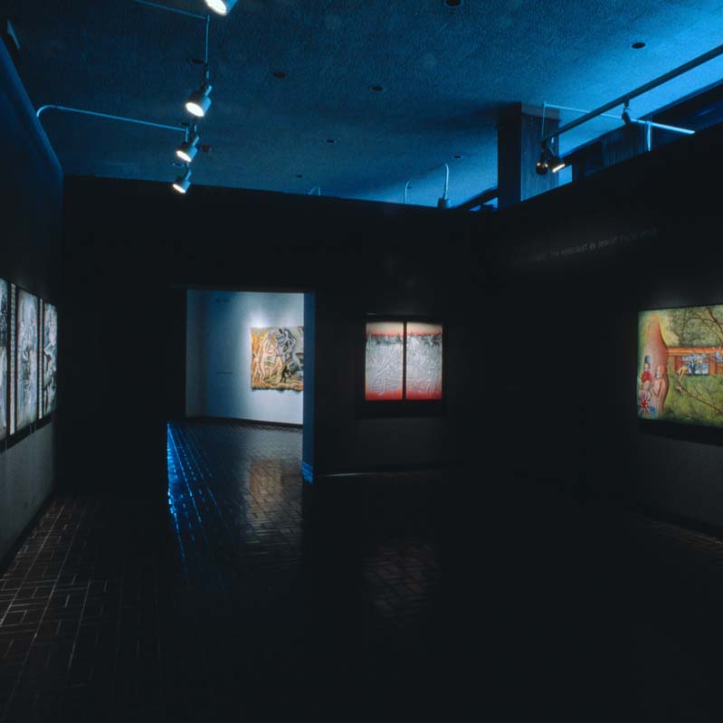 Color photograph of artworks on the walls of a darkened gallery