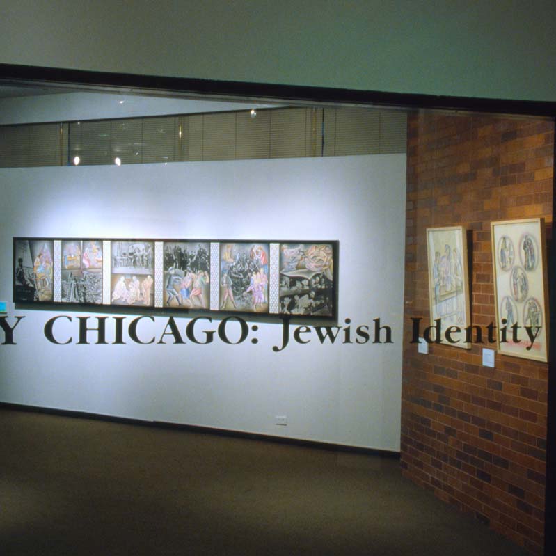 Color photograph of artworks installed on white and brick walls, seen through a window with text on it
