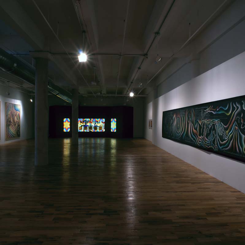 Color photograph of paintings and a stained glass work in a darkened gallery with white walls
