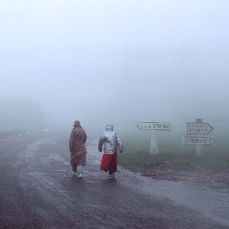Color photograph of two figures walking away on a road in the fog
