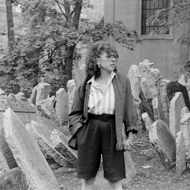 Black-and-white photograph of Judy Chicago standing in front of fallen and crooked gravestones