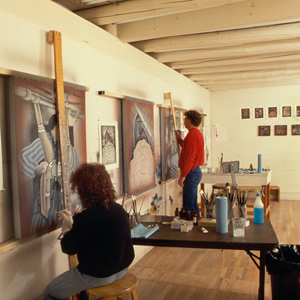Color photograph of Judy Chicago (seated) and Donald Woodman painting works on wall-mounted easels in an art studio