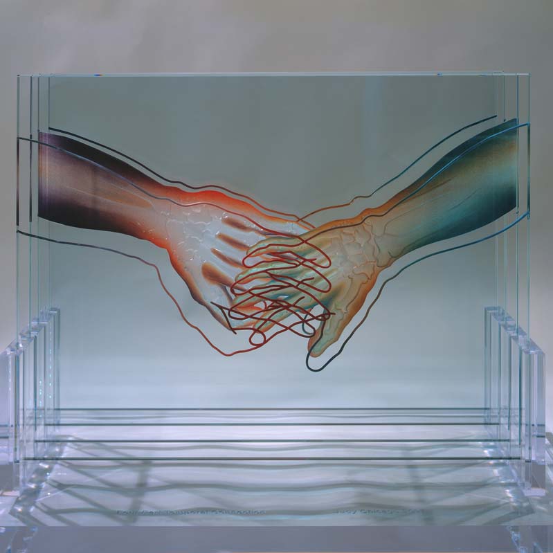 Sculpture of two arms holding hands painted in multiple colors on four layered clear rectangles on a clear plinth where one of the arms has exposed bones