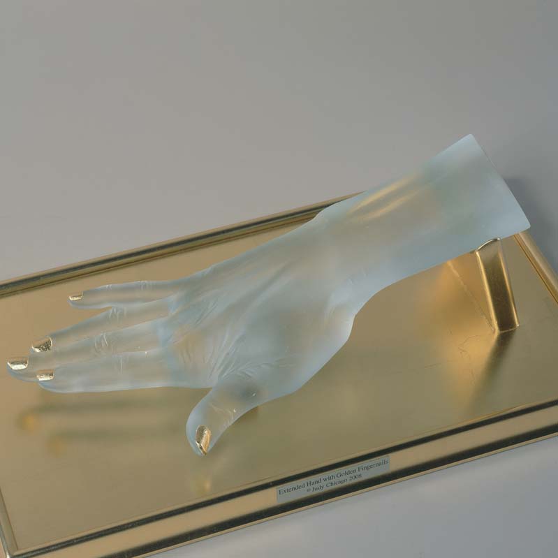 Sculpture of a translucent white hand with fingers splayed and gold fingernails on a gold plinth