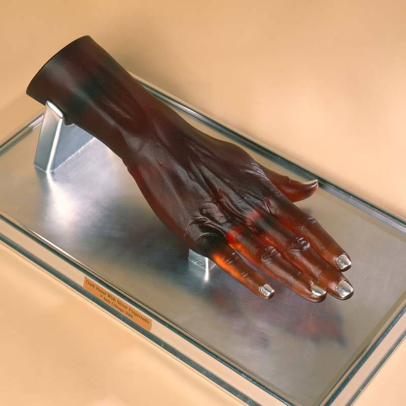 Sculpture of a translucent brown hand with silver fingernails on a clear plinth