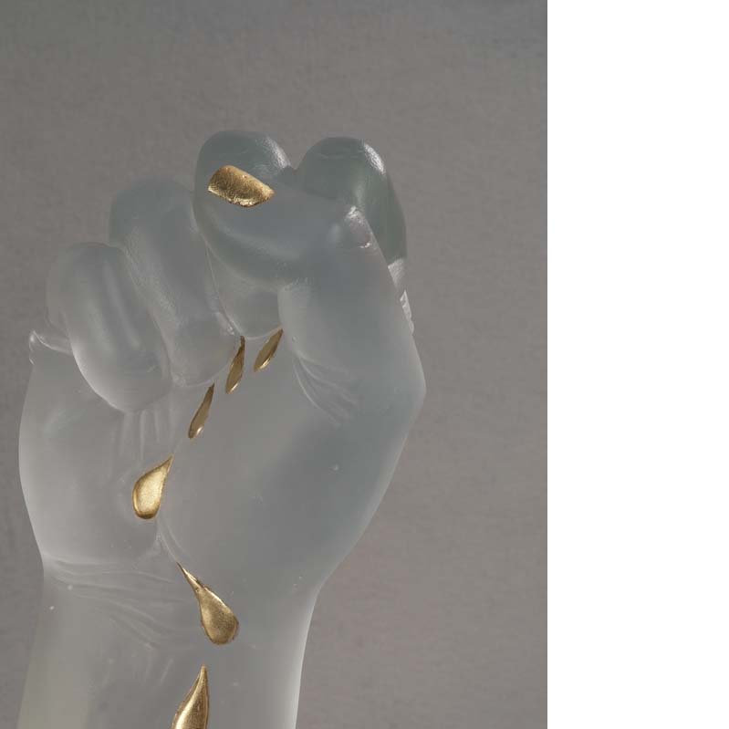 Detail of a sculpture of a translucent white raised fist with gold tears trailing down the arm