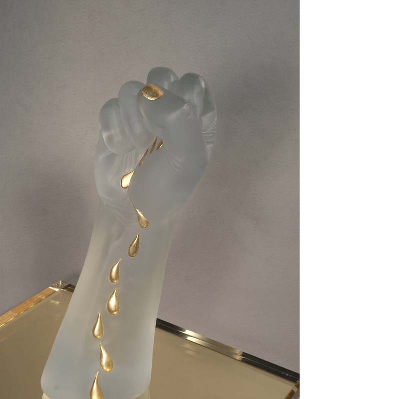 Sculpture of a translucent white raised fist with gold tears trailing down the arm on a gold plinth