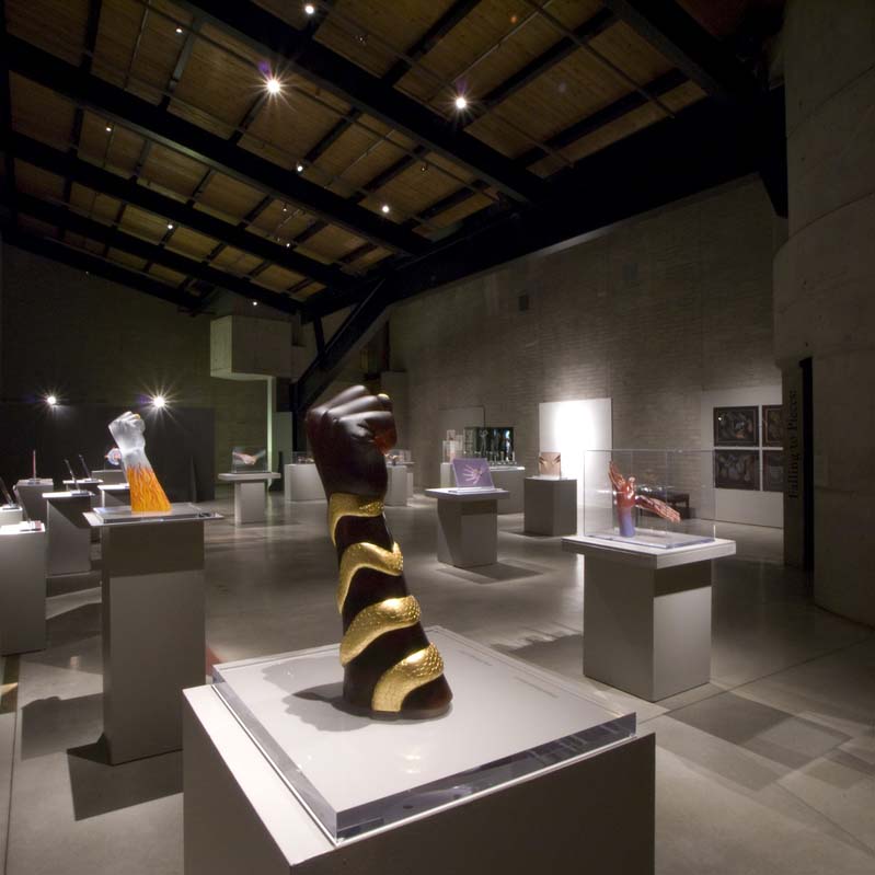 Color photograph of a gallery installation with multiple sculptures on gray pedestals and several works installed in a large, semi-darkened room with gray walls and a pitched ceiling