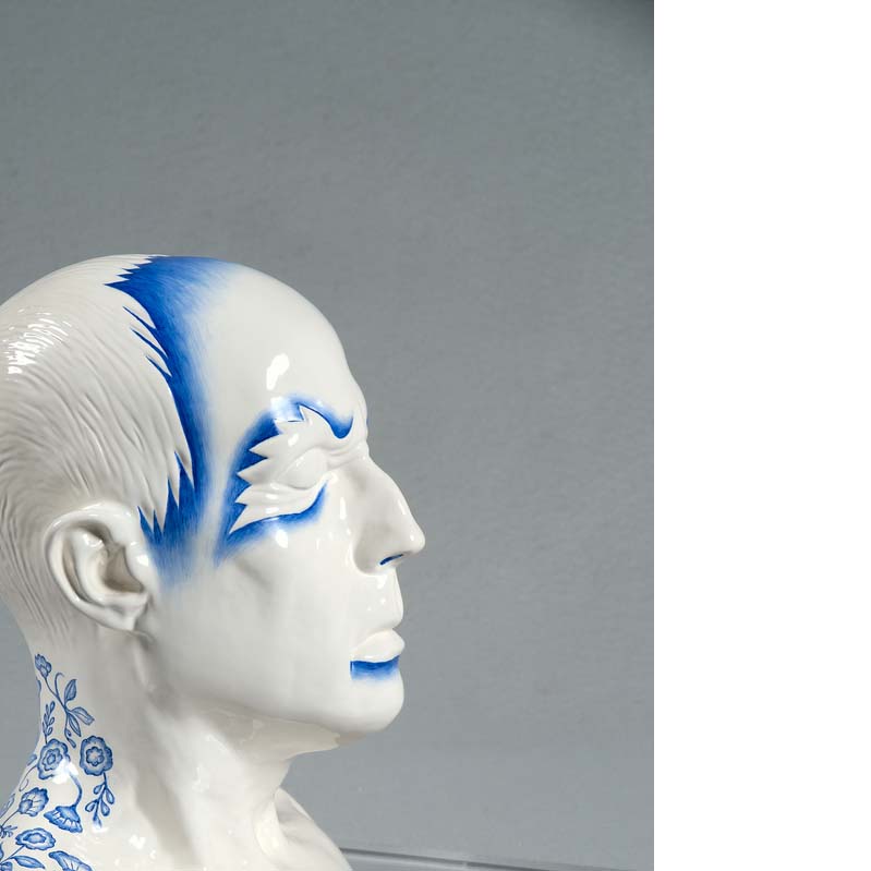 White and blue sculpture of a man's head with blue flame-like shapes around his eyes and flowers on his neck on a clear plinth