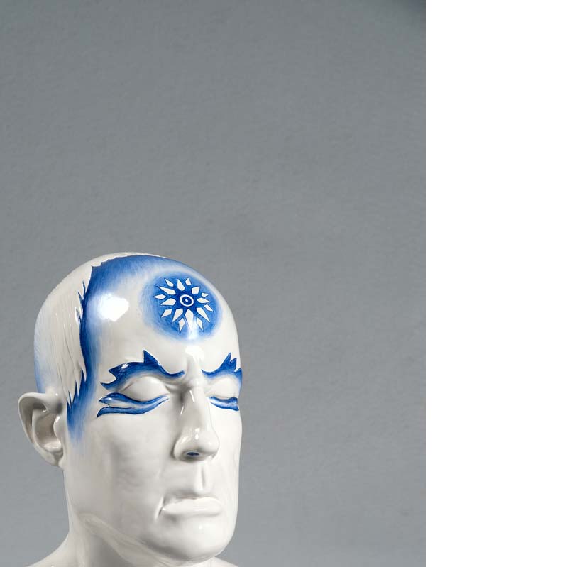 White and blue sculpture of a man's head with blue flame-like shapes around his eyes and a flower on his forehead on a clear plinth