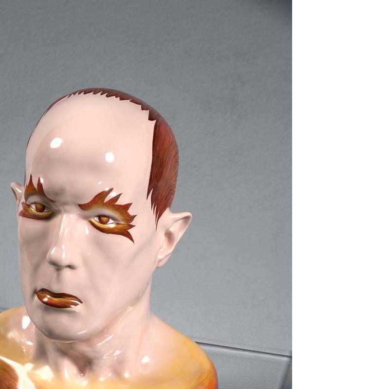 White, brown, and orange sculpture of a man's head with brown flame-like shapes around his eyes on a clear plinth