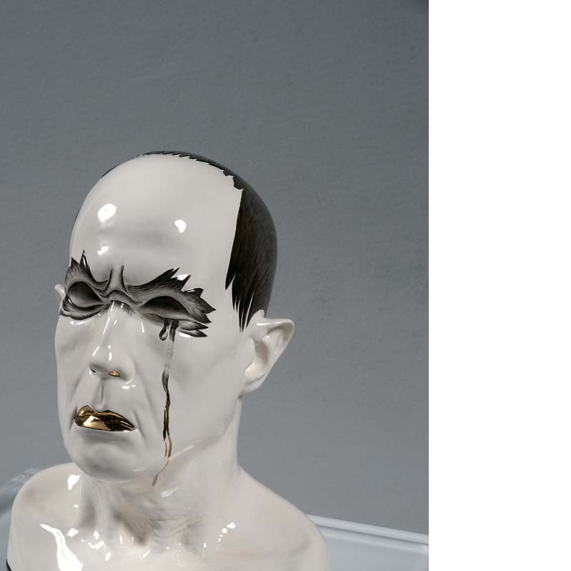 Sculpture in white, black, gray and gold of a head of a crying man with flame-like shapes around his eyes on a clear plinth