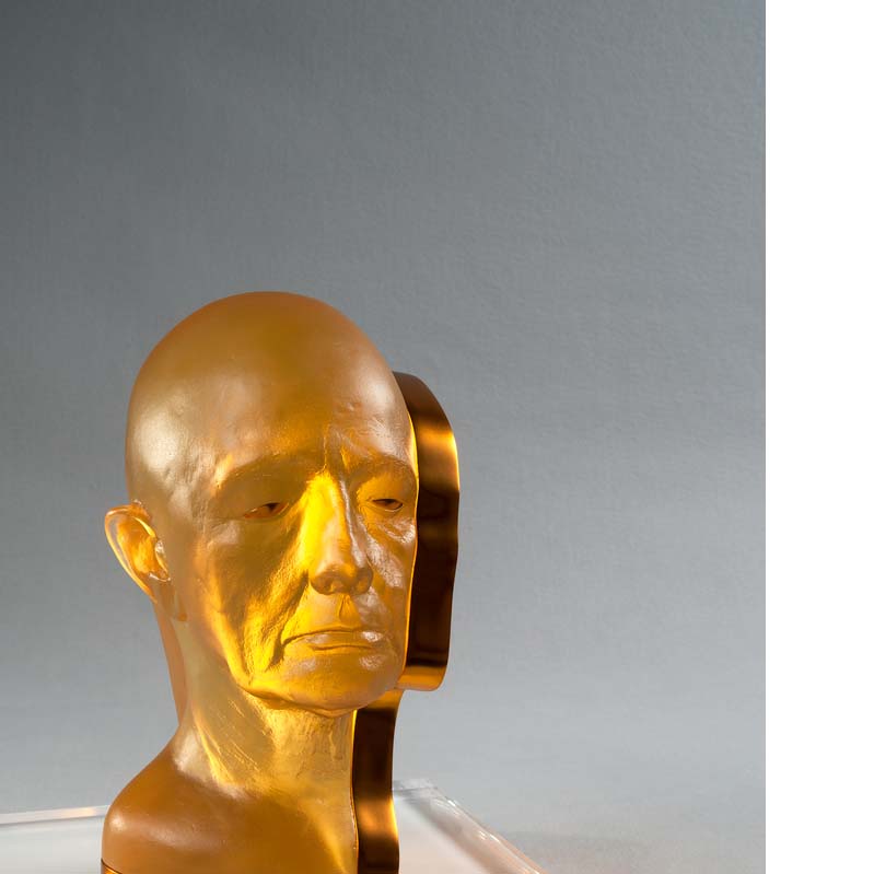 Translucent yellow sculpture of a bald head with a shiny gold strip along the base and one side on a white plinth