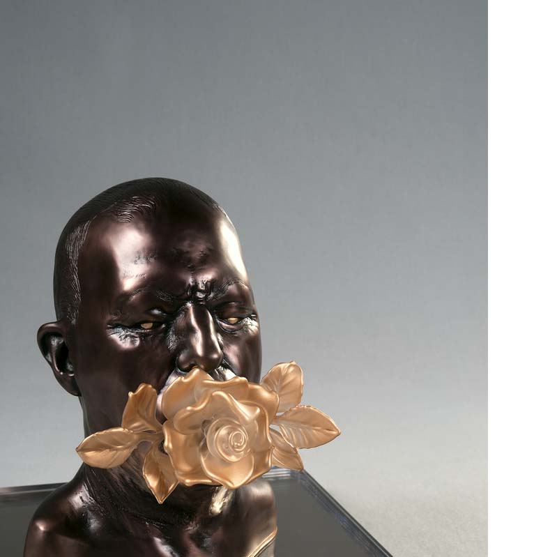 Shiny brown sculpture of a bald head with a gold flower in its mouth on a mirrored plinth