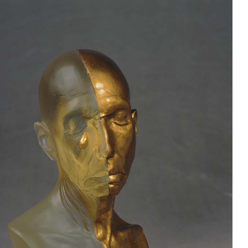 Sculpture of a bald head crying divided vertically in half where one half is translucent yellow and the other is metallic gold