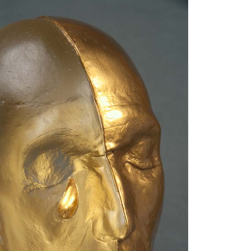 Detail of a sculpture of a bald head crying divided vertically in half where one half is translucent yellow and the other is metallic gold
