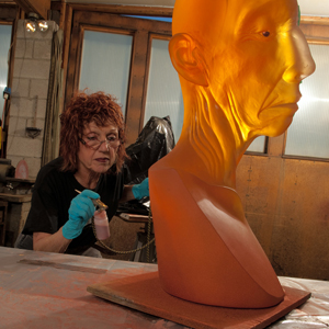Color photograph of a sculpture of half of a translucent orange bald head on a table Judy Chicago is working on another sculpture in the background