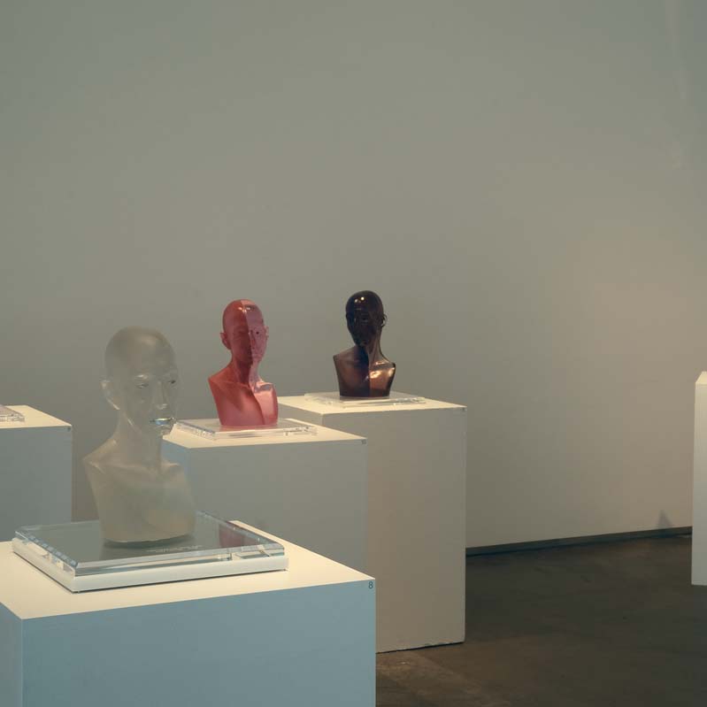 Color photograph of five bald head sculptures each on a white pedestal in a room with white walls
