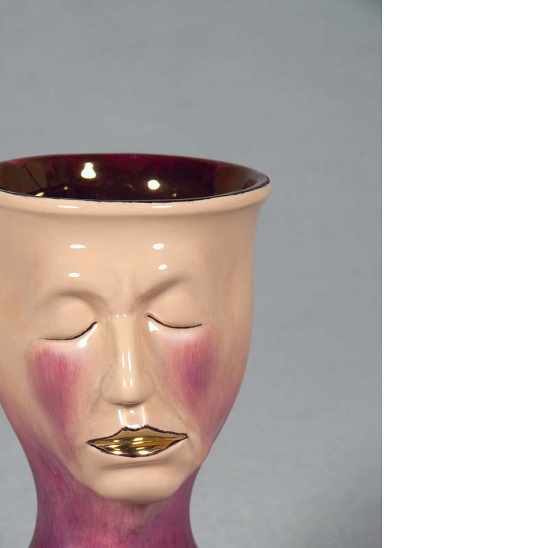 Mug in shades of beige, purple, and gold with a face atop a flared base The face's eyes are closed