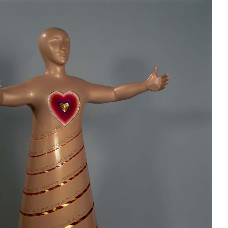 Gold sculpture of a figure encircled with gold text and spiraling lines with outstretched arms and a red and gold heart affixed to its chest